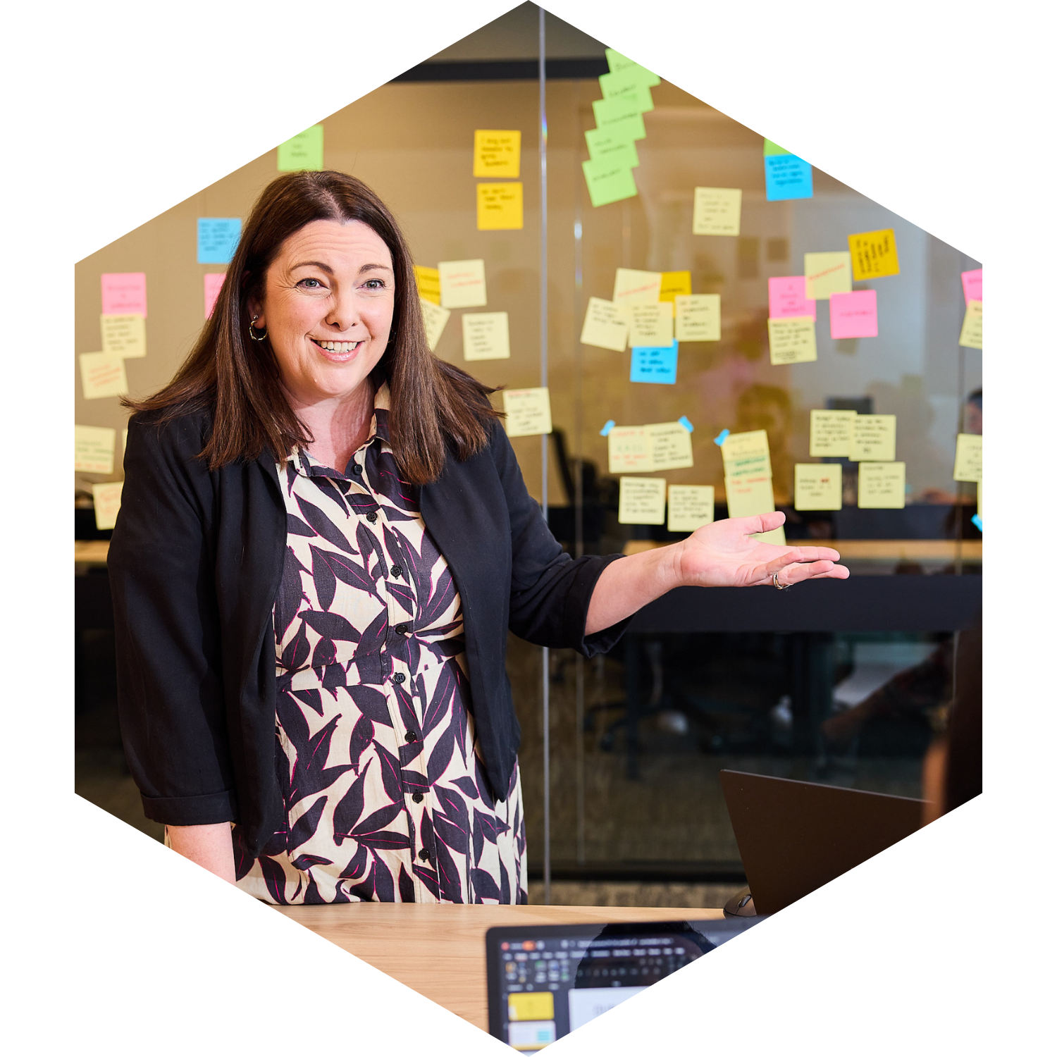 A woman standing in front of a glass wall covered with colorful sticky notes, gesturing towards it with a smile.