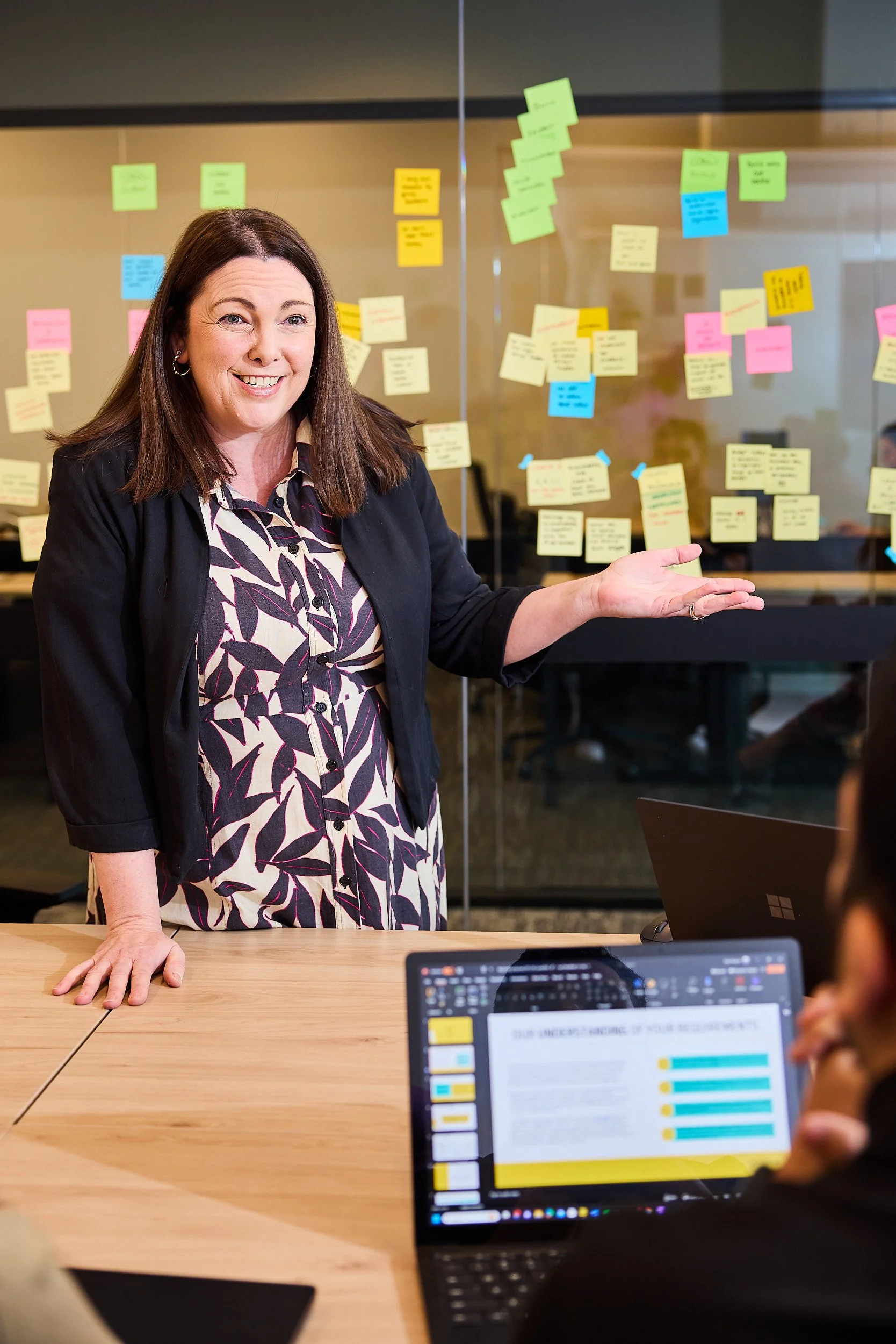 A woman smiling and gesturing with her right hand in a meeting room with a glass wall covered in colorful sticky notes. Laptops are on the table, showing presentations and notes.