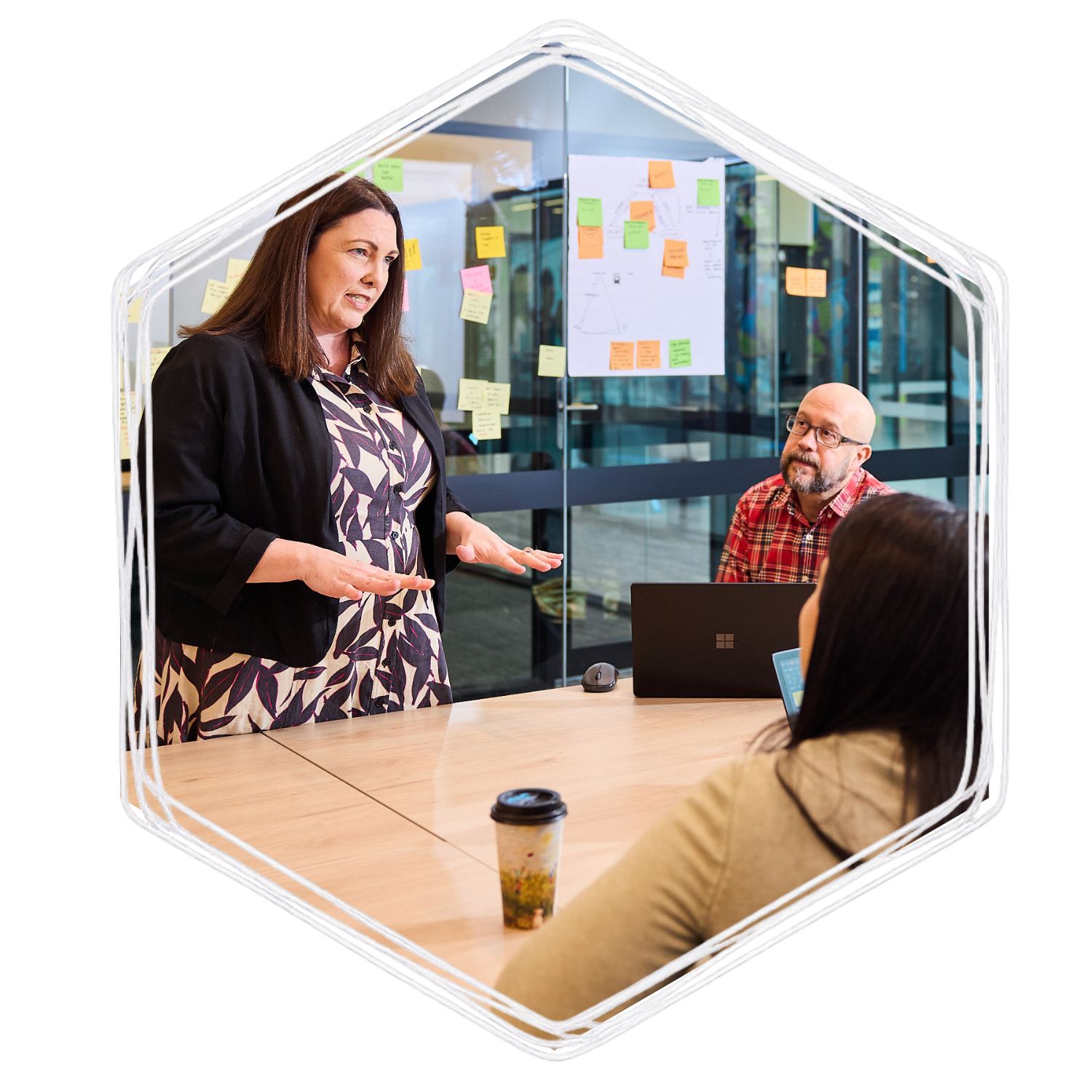 A woman stands at a conference table, speaking to a group of seated people in an office meeting room with colorful sticky notes on the glass walls.