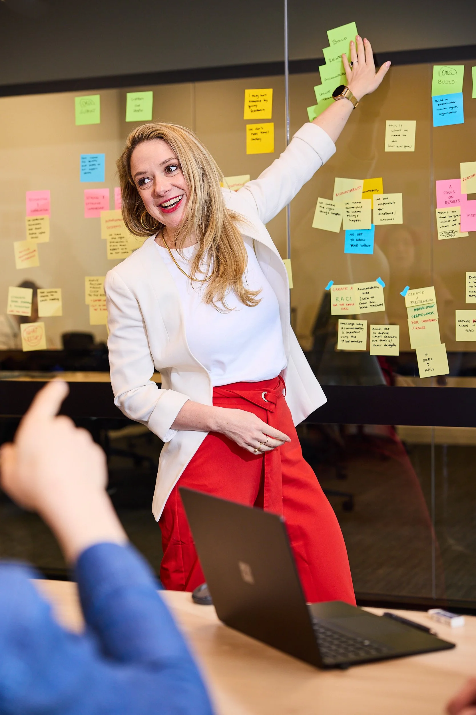 A woman in a white blazer and red pants is smiling and gesturing towards a glass wall covered with colorful sticky notes in a meeting room. In the foreground, there is a laptop and a person's hand pointing towards the woman.