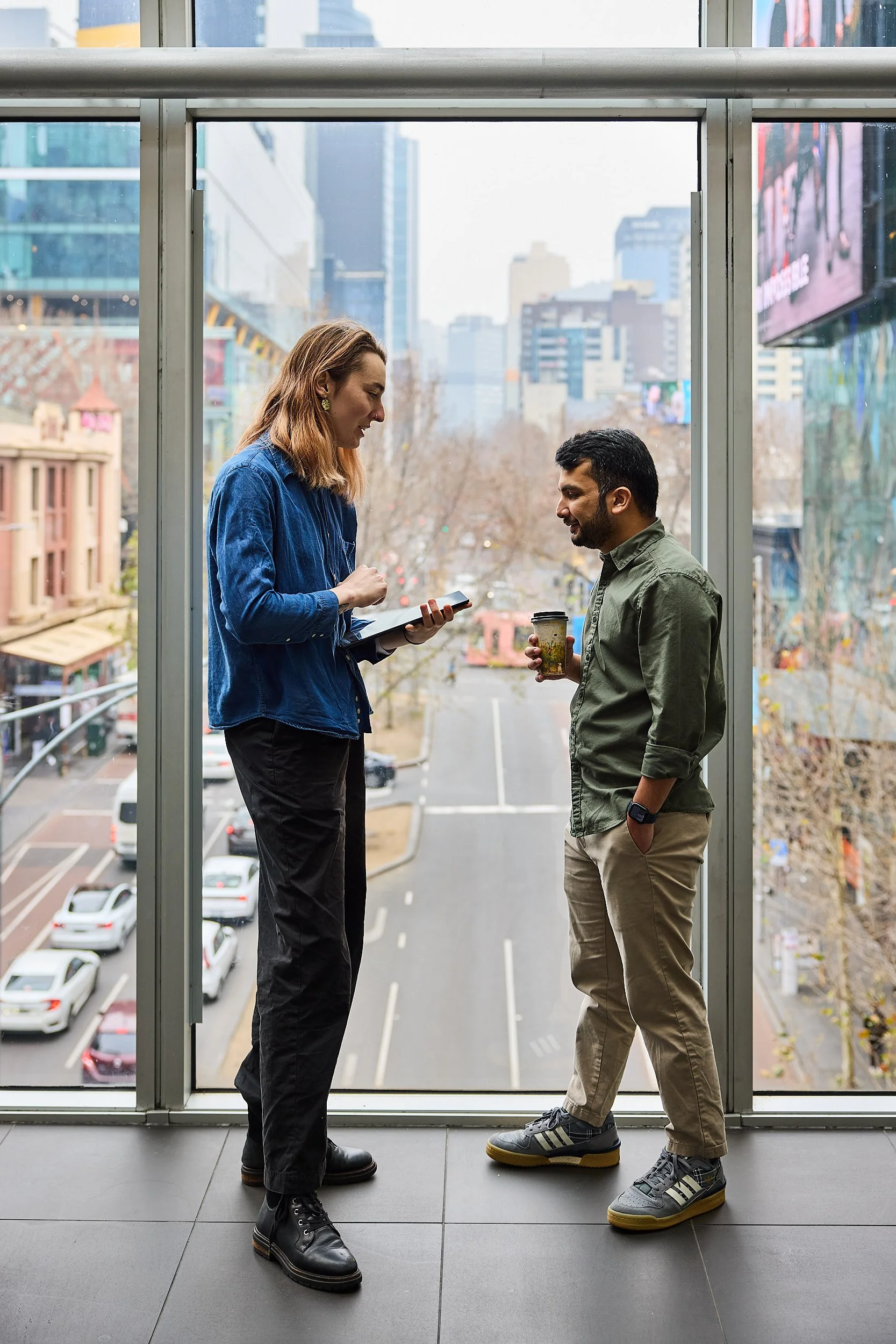 Two people talking inside a building near a large window with a city view. The woman on the left is holding a notepad and pen, while the man on the right is holding a coffee cup. They are casually dressed, and the cityscape with tall buildings and streets is visible outside.