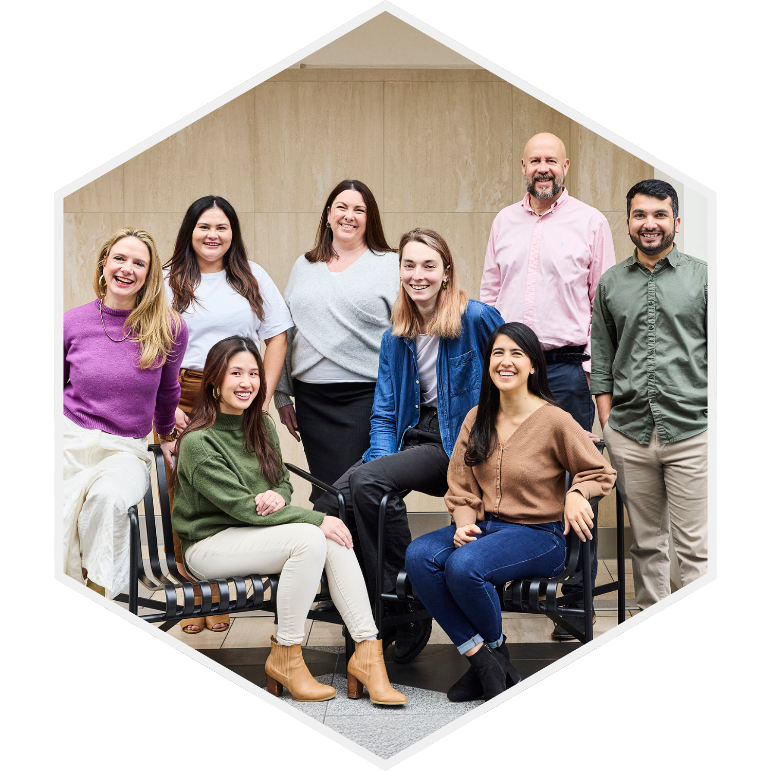 Group of nine diverse people smiling and posing for a photo in an indoor setting with beige walls, some seated and some standing.