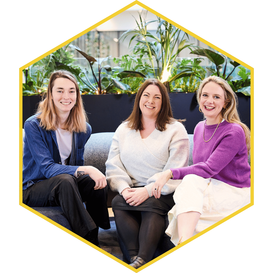 Three women sitting on a sofa in front of a leafy indoor garden, smiling at the camera.