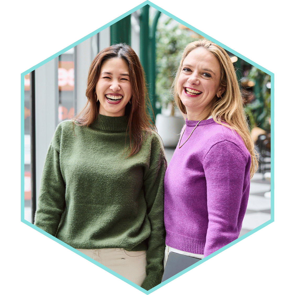 Two women smiling and laughing outdoors during daytime, standing close together near glass windows and greenery, wearing colorful sweaters.