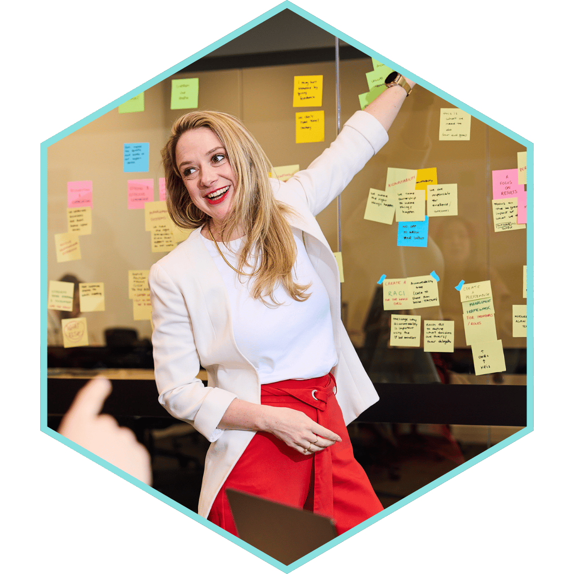 Woman in a white blazer and red skirt giving a presentation with colorful sticky notes on a glass wall.