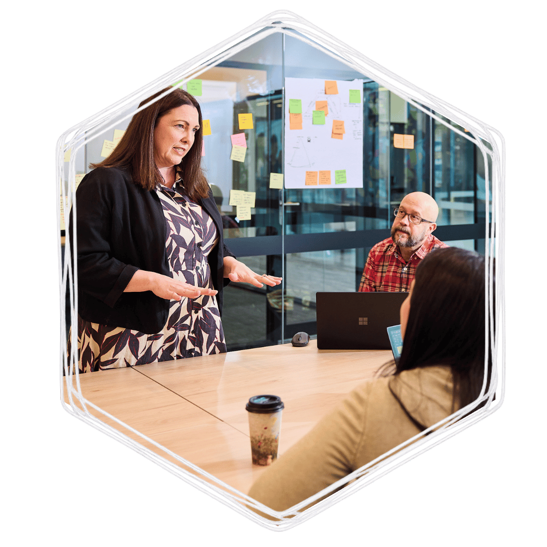 A woman standing and speaking to two seated women in a modern office with glass walls and sticky notes on the wall in the background.