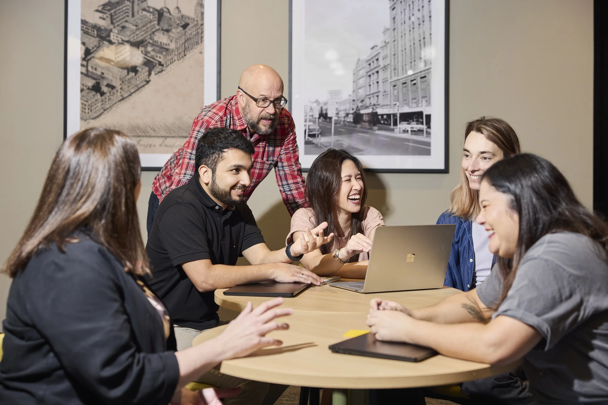 Group of six people sitting around a table with laptops, engaging in a lively discussion, with a man standing and leaning over while speaking, in a room with framed black-and-white cityscape photographs on the wall.