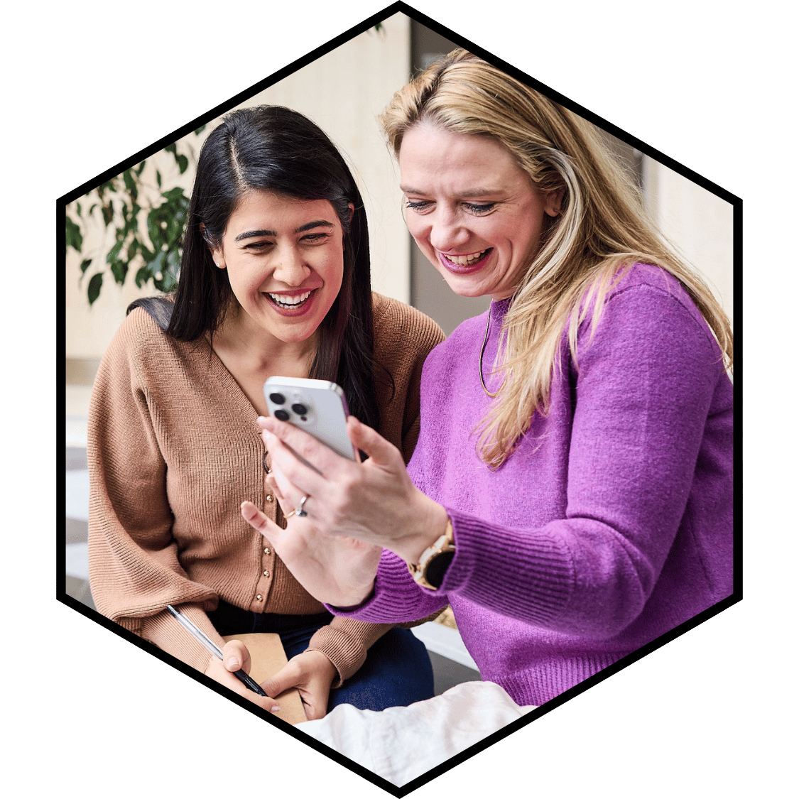 Two women laughing while looking at a smartphone together, sitting indoors.