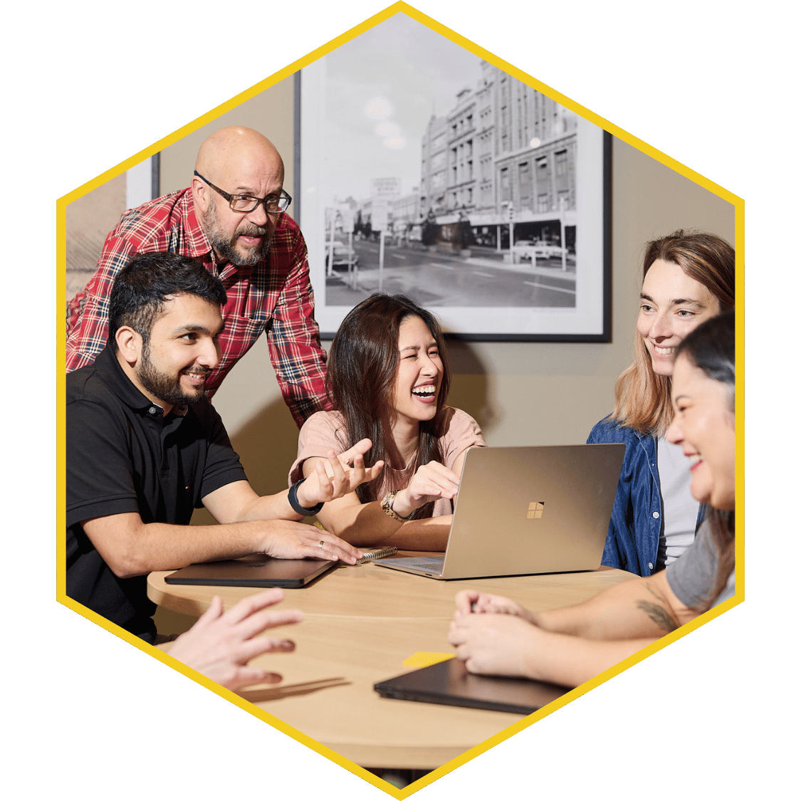 Group of people sharing a laugh around a laptop during a meeting in a conference room with a black and white cityscape photograph on the wall.