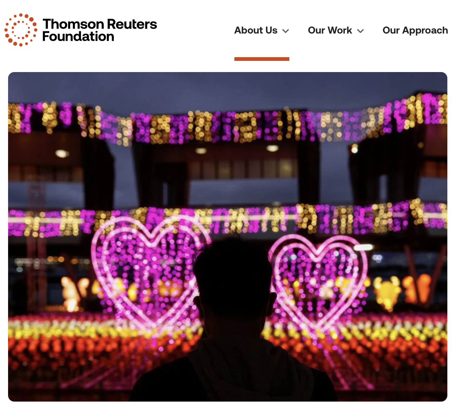Person standing in front of colorful heart-shaped neon lights and decorative hanging string lights during evening.