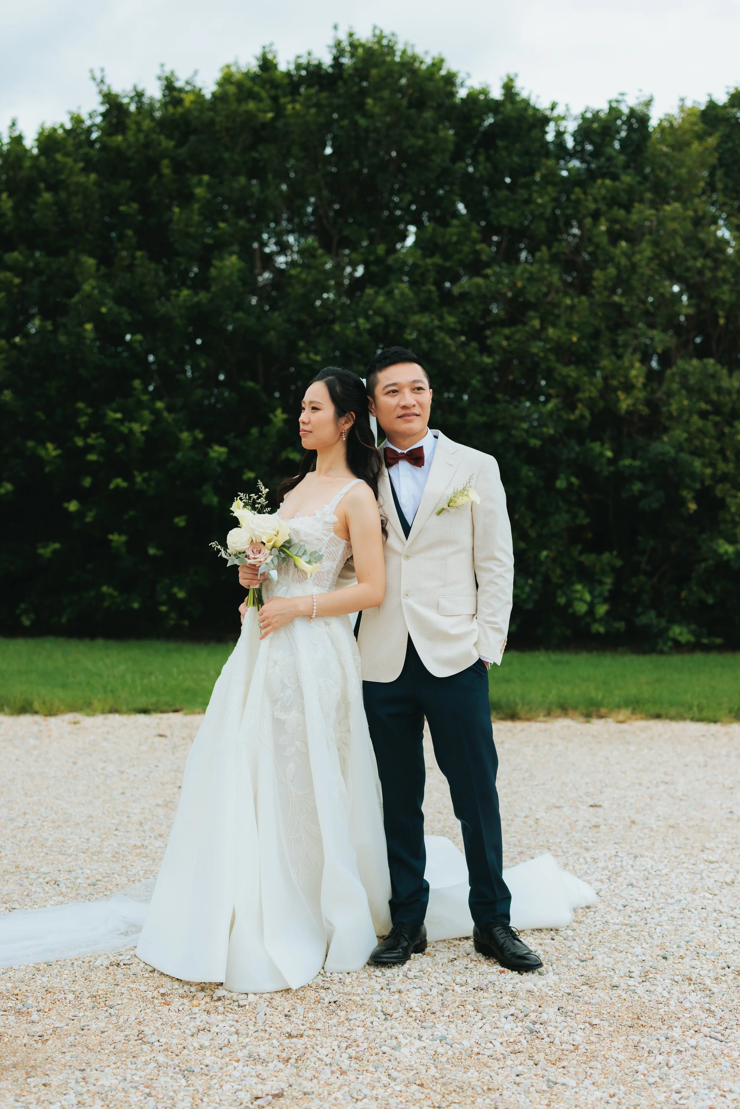 A man in a black tuxedo with a bow tie stands behind a woman in a white strapless gown outside a building with stone walls and steps.
