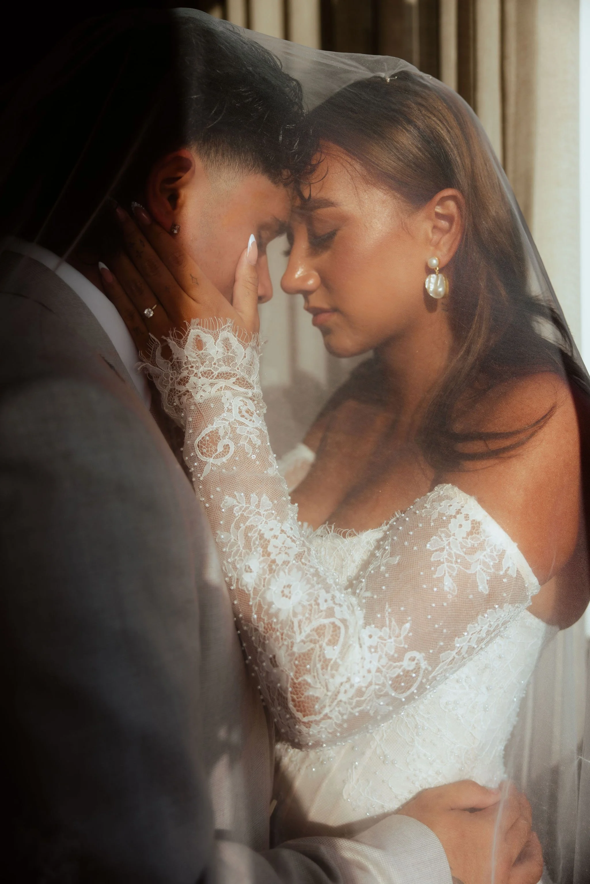 Bride hugging groom against brick wall, smiling with eyes closed, wearing a white lace wedding dress and veil.