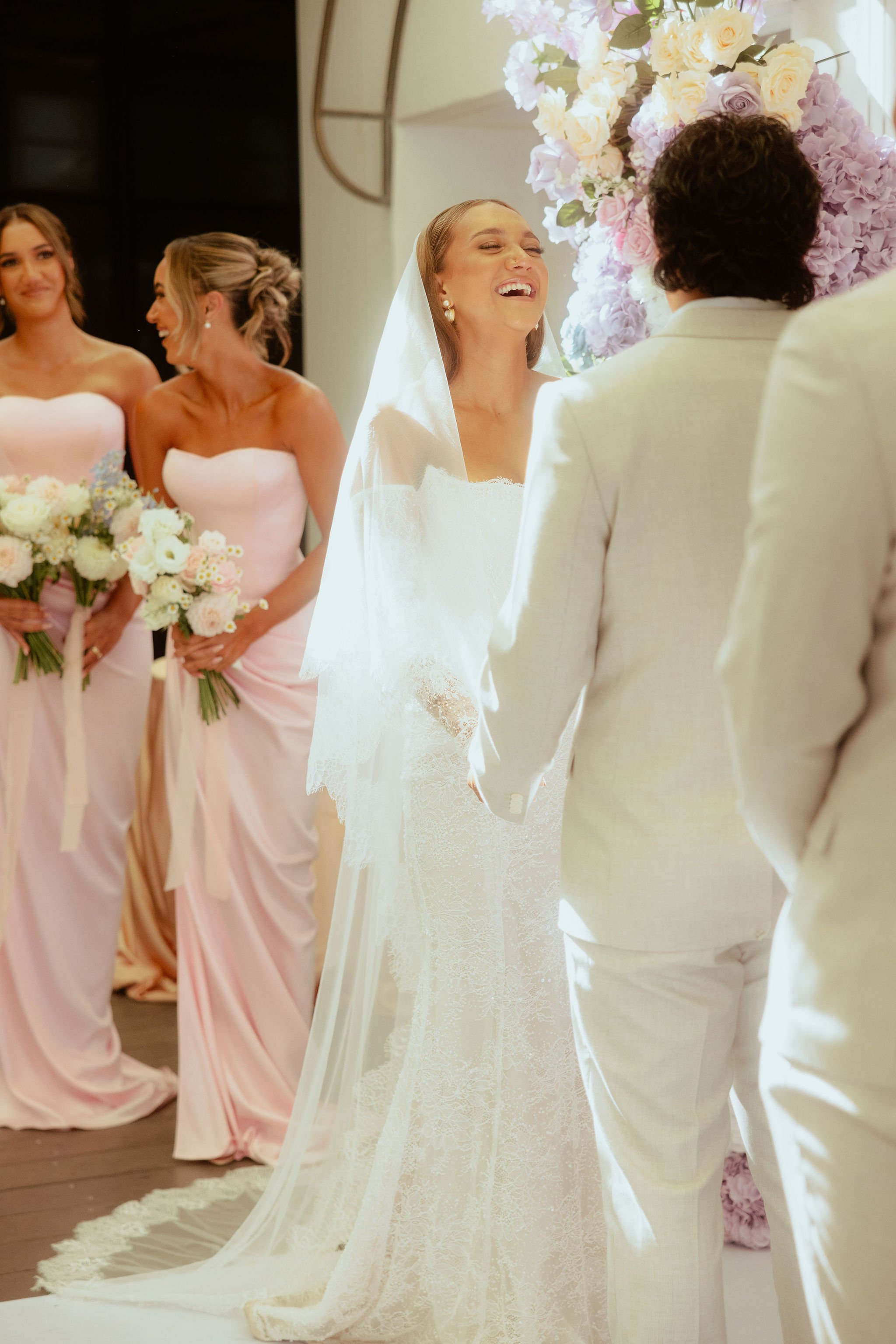 Bride hugging groom against brick wall, smiling with eyes closed, wearing a white lace wedding dress and veil.