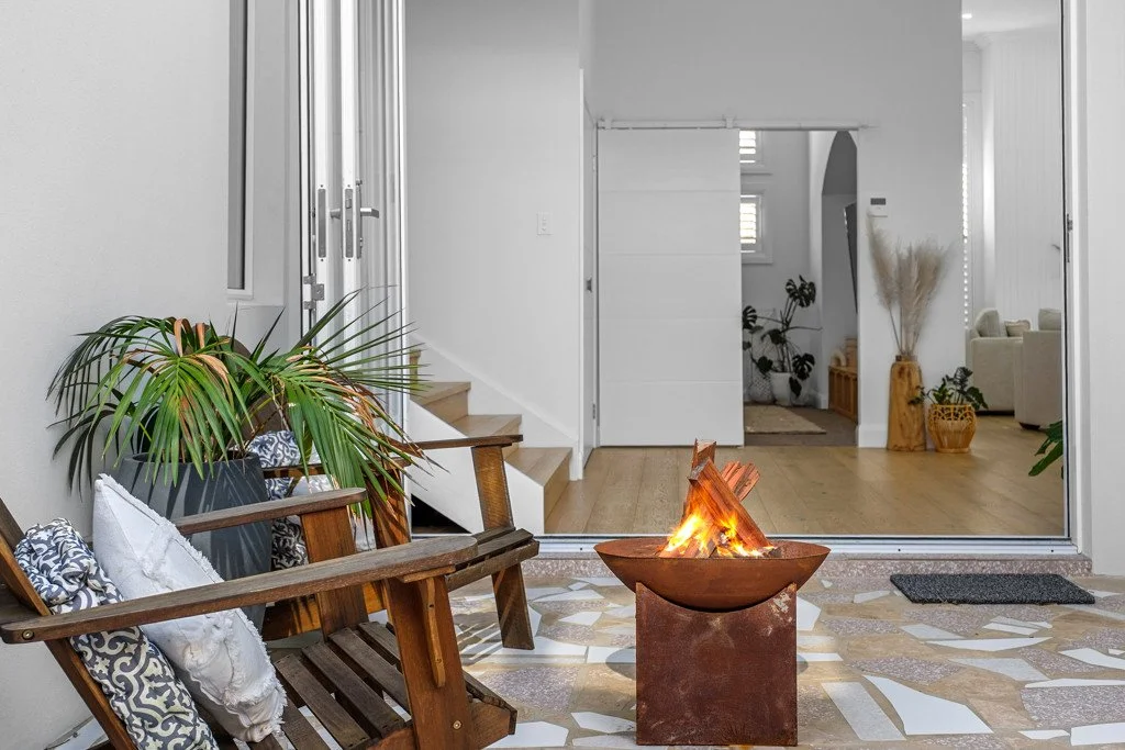 Outdoor patio with wooden benches, decorative pillows, a fire pit, and potted plants, leading into a bright, modern home interior.