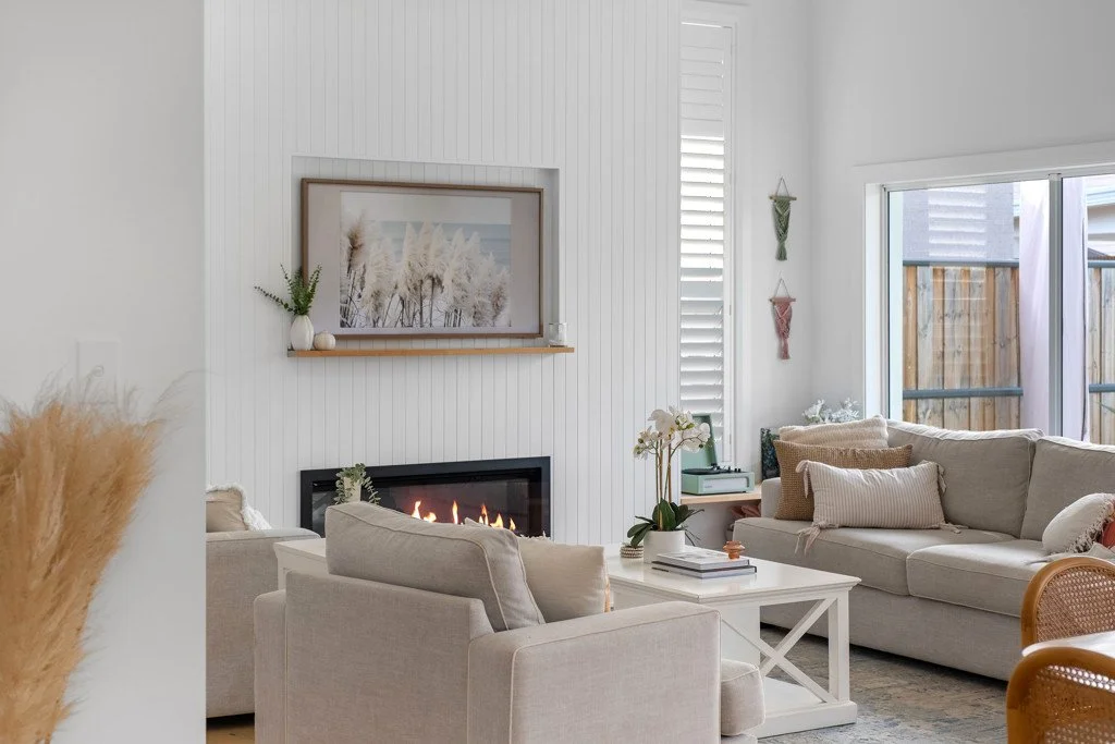 A cozy living room with a white fireplace, beige sofas, a coffee table with books and a plant, and a large window with view of a wooden fence outside.