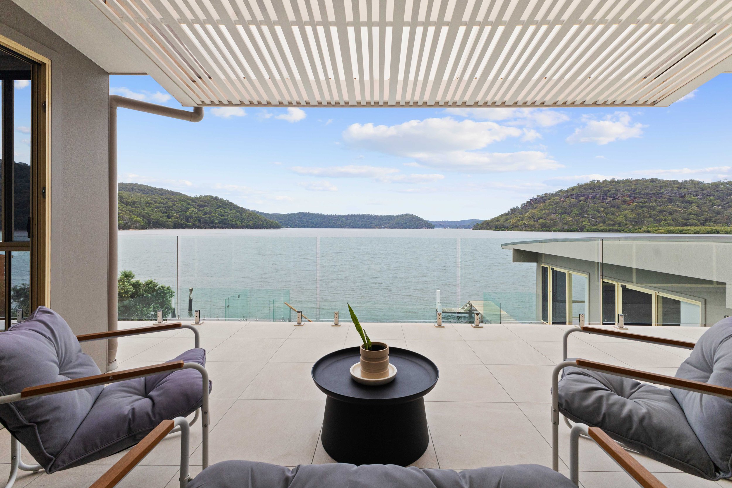 Balcony with glass railing overlooking a lake and green hills, with patio furniture and a small potted plant on a black side table.
