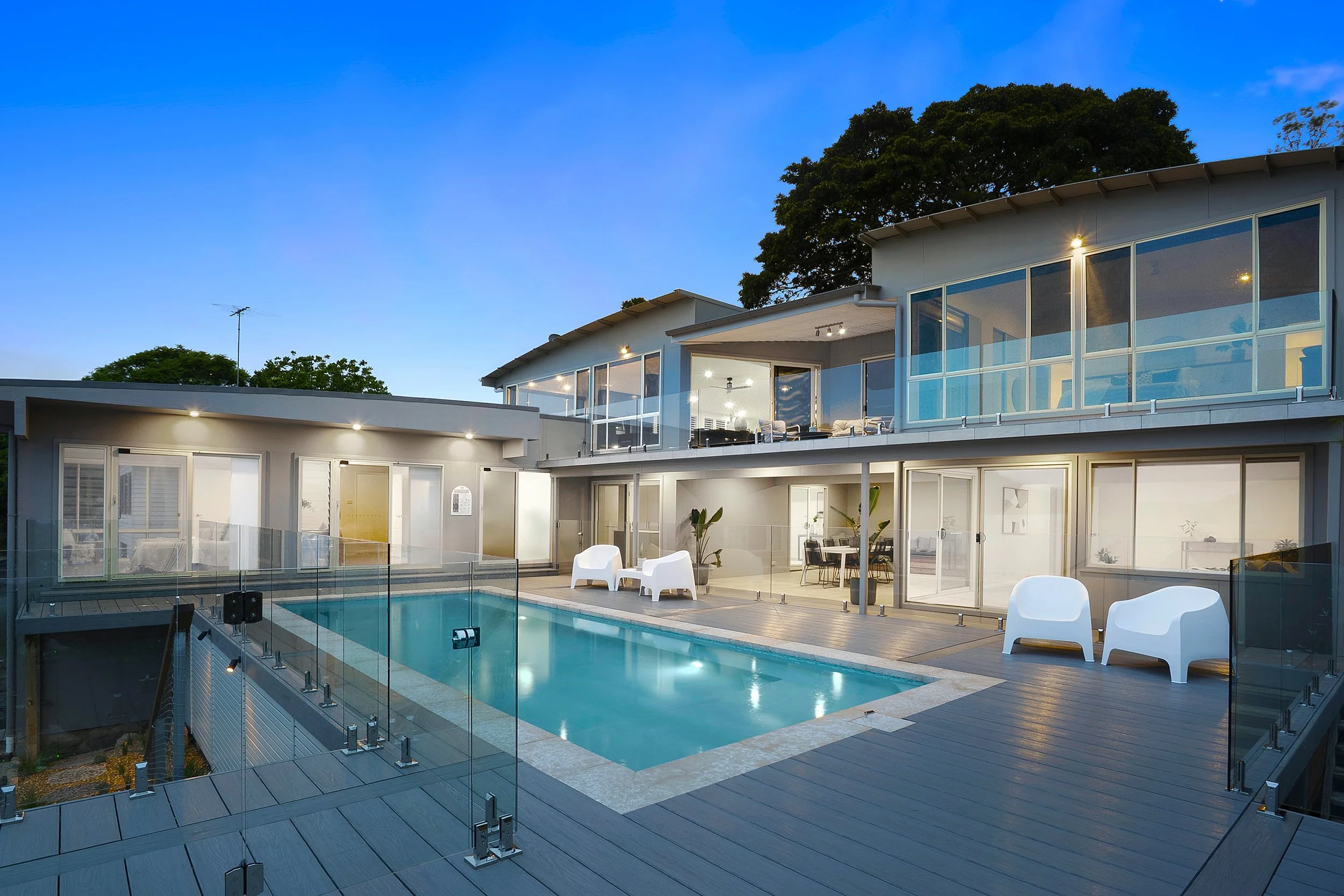 Modern house with large glass windows, balcony, and a swimming pool with a glass fence in the backyard during evening.