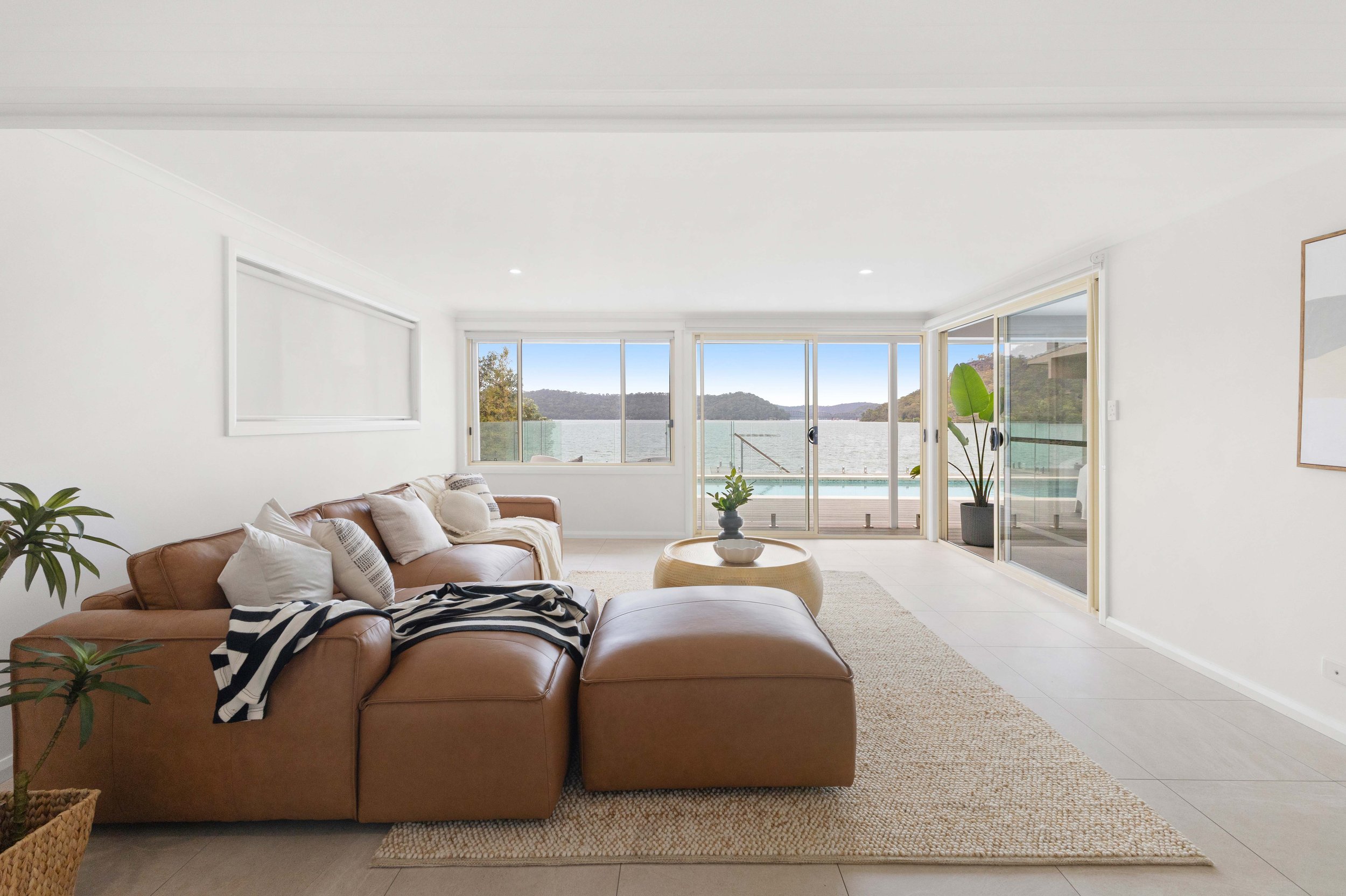 Living room with large windows and sliding glass doors overlooking a body of water and hills, furnished with a tan leather sectional sofa, an ottoman, potted plants, and minimal decor.