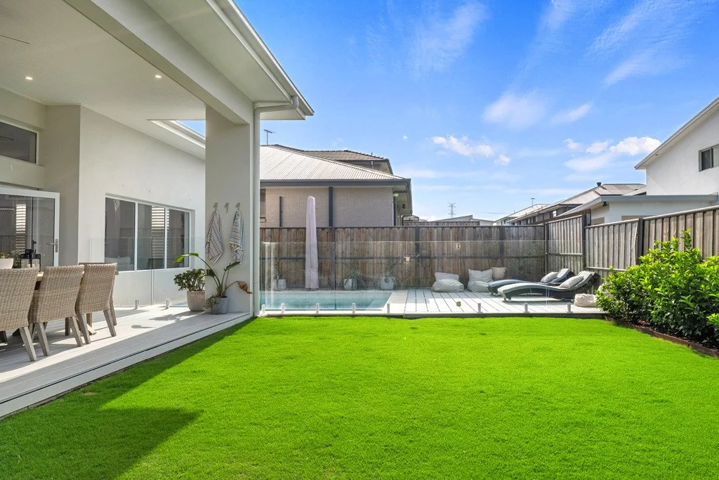A backyard with a green lawn, patio furniture, and a swimming pool, enclosed by a wooden fence under a bright blue sky with some clouds.