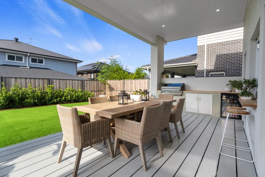 Outdoor covered patio with a wooden dining table and six wicker chairs, outdoor kitchen with grill and cabinets, green lawn, garden fence, and neighboring houses in the background.