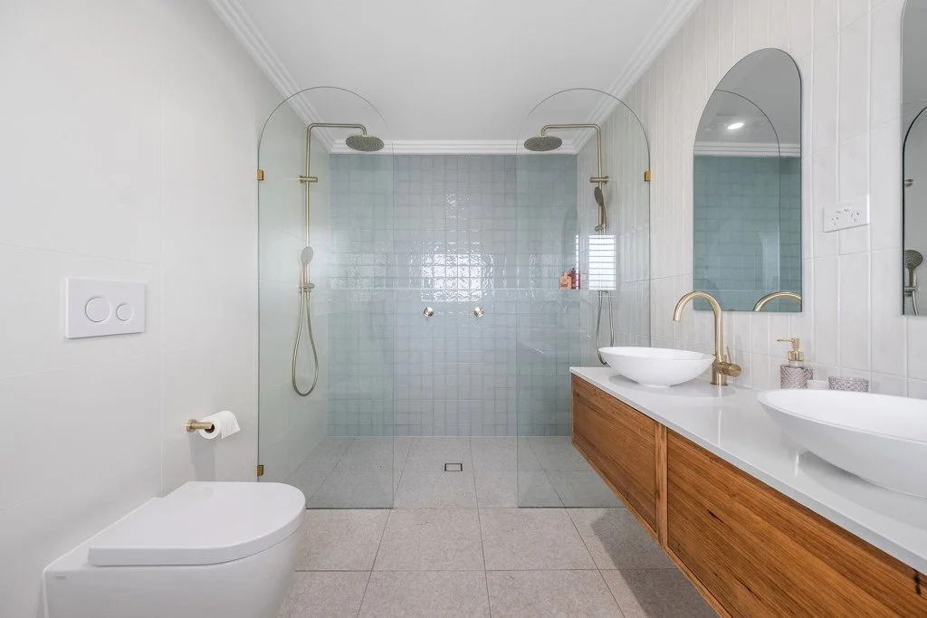 Modern bathroom with a glass-enclosed shower, two vessel sinks with gold fixtures, wooden vanity, and a toilet.