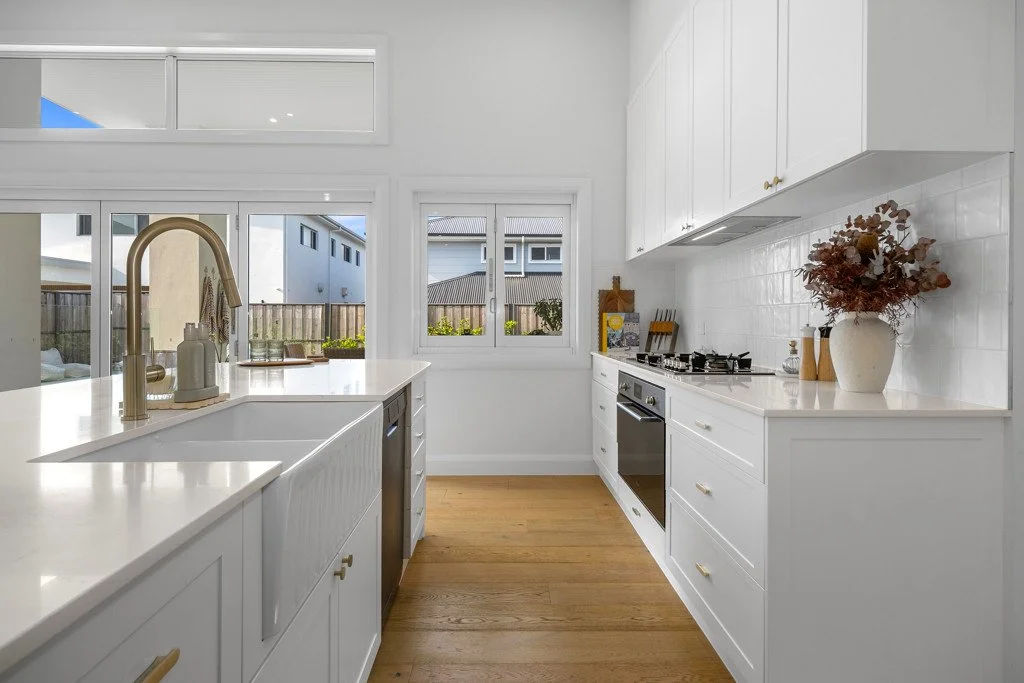 Modern kitchen with white cabinets, a white countertop, a farmhouse sink, a built-in oven, gas stove, and decorative items including a plant in a vase and kitchen utensils, with large windows providing natural light.
