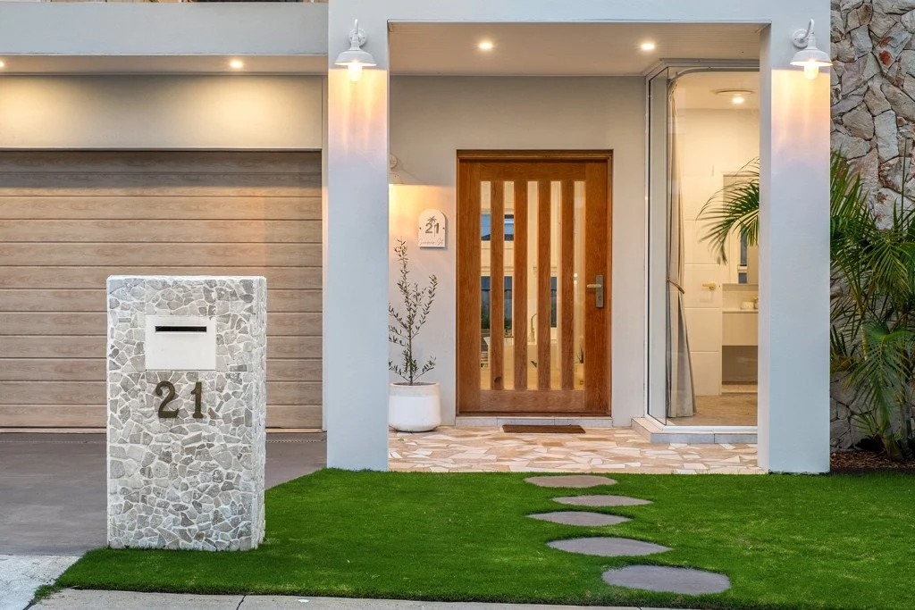 The image shows the front entrance of a modern house with a stone and wood design. There is a mailbox with the number 21 on a stone stand, a pathway with stepping stones on the grass, and a wooden door. The house has exterior lighting and potted plan