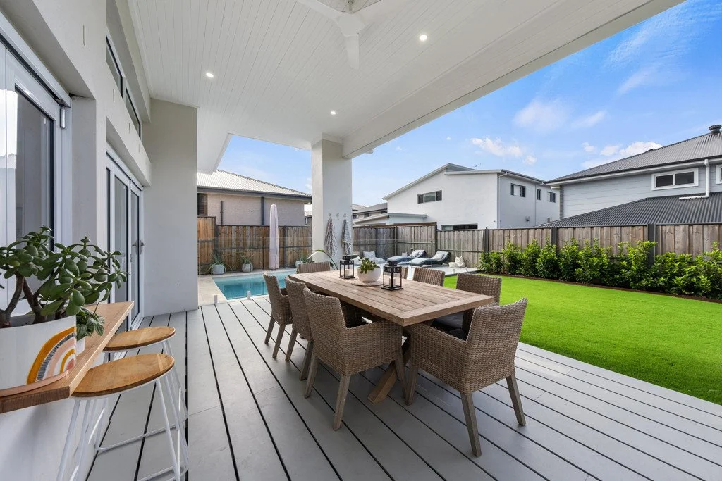 Backyard patio with dining table and wicker chairs, a little swimming pool, lounge chairs, lush green lawn, and neighboring houses in the background.