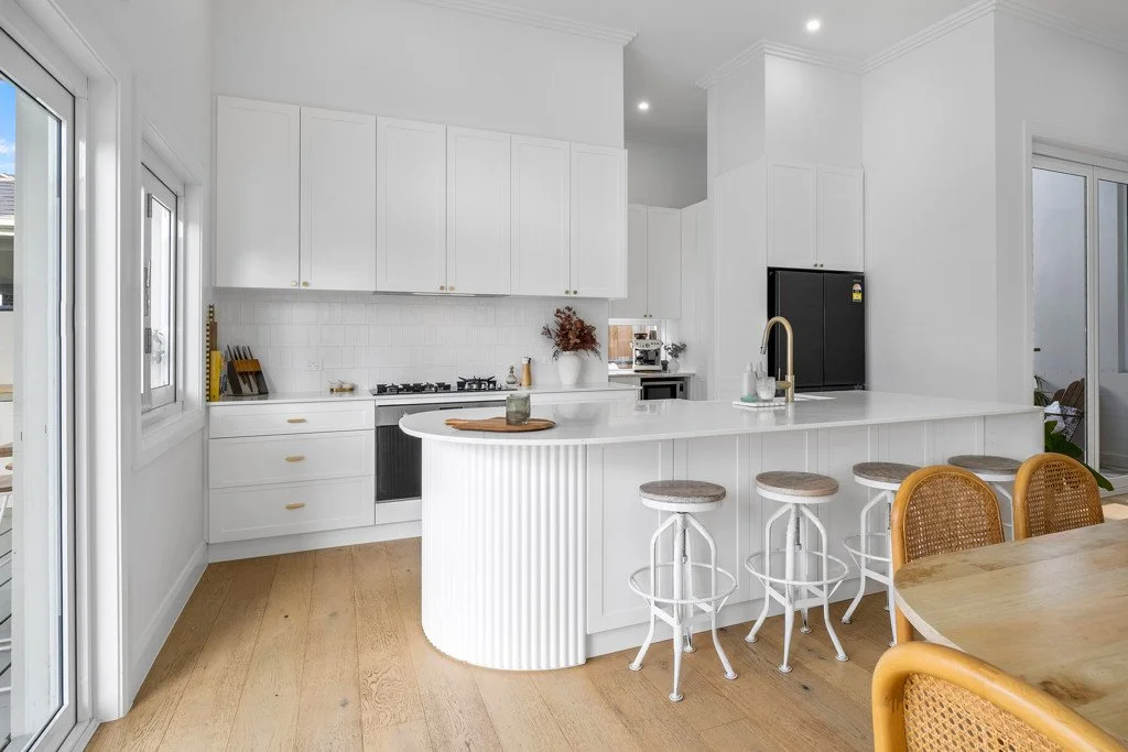 Modern white kitchen with a breakfast bar, surrounded by four white stools with woven seats, and a wooden dining table with yellow-caned chairs. Large windows and sliding glass doors let in natural light.