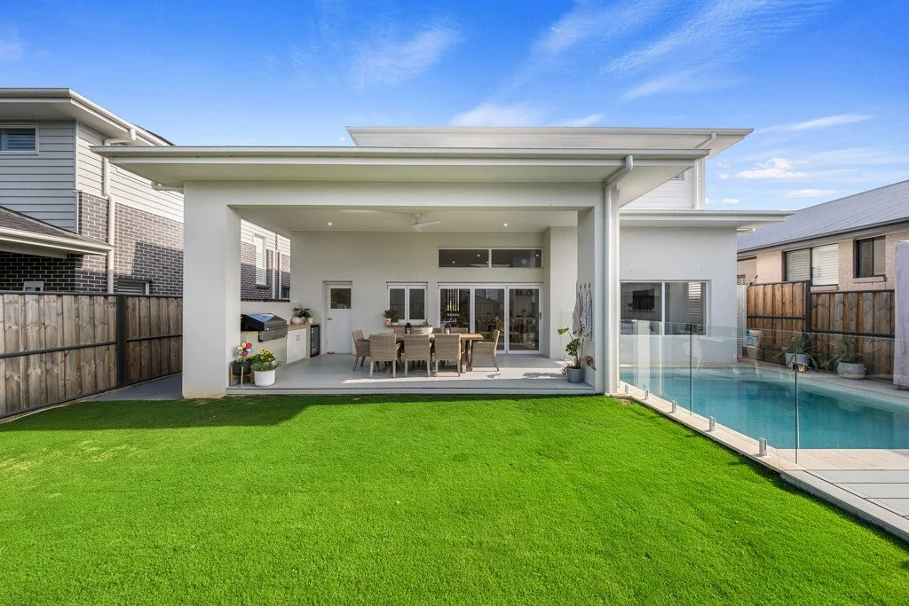 Modern backyard with a covered patio, outdoor dining table, and a swimming pool with glass fencing.