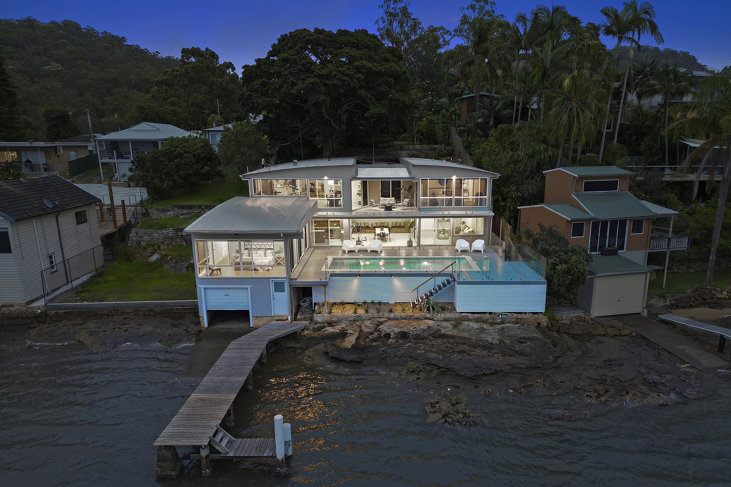 A modern multi-story house located on a waterfront, with a dock extending over the water, surrounded by trees and neighboring houses.