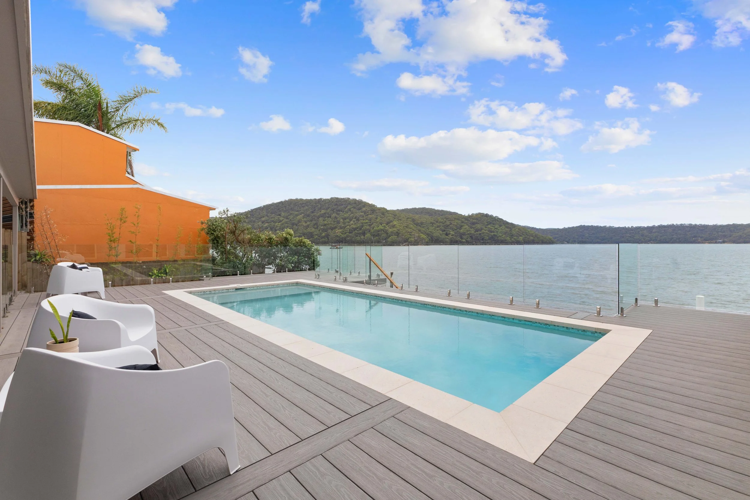 Outdoor pool on a wooden deck overlooking a body of water with green hills in the distance, clear blue sky, white clouds, and modern white chairs with black cushions and a small potted plant on the deck.
