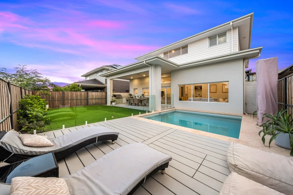 Modern backyard with pool, outdoor lounge chairs, grill area, green lawn, and a house with large windows at sunset.