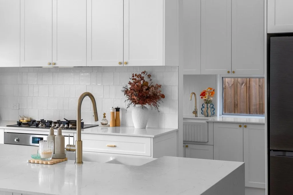 Modern white kitchen with gold hardware, gold faucet, white cabinets, white marble countertops, window with wooden fence outside, and decorative vases with flowers.