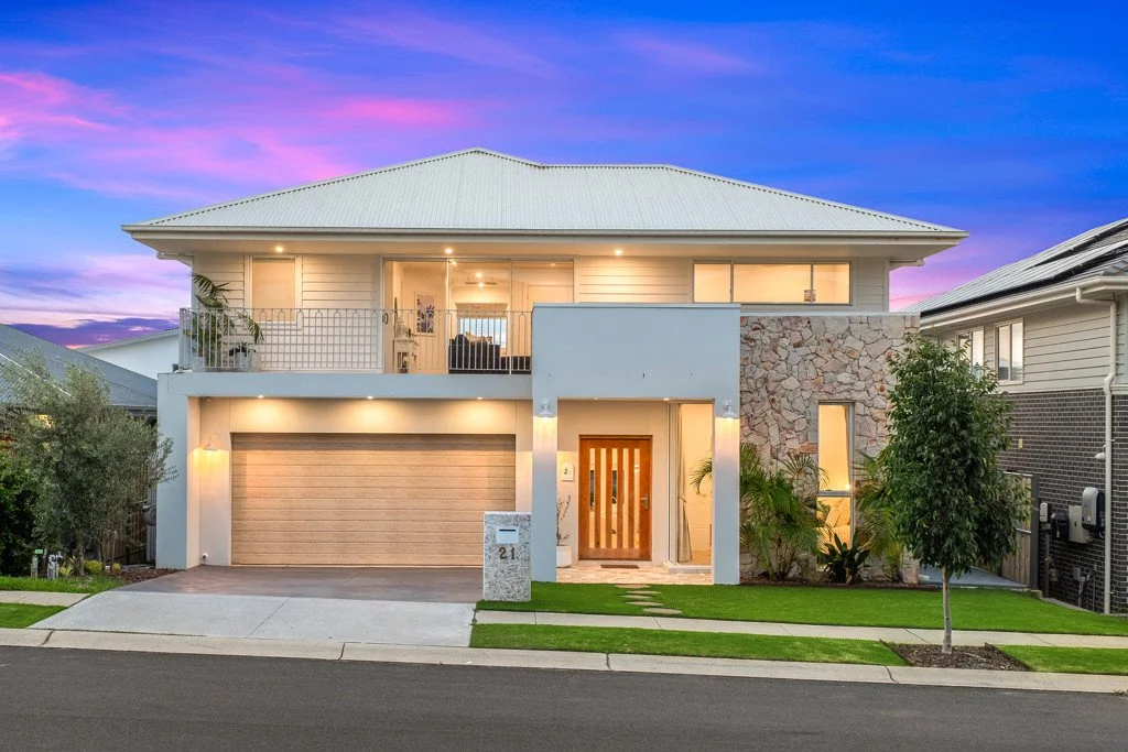 Modern two-story house with a beige garage door, wooden front door, and a small front yard with grass and trees, under a colorful sunset sky.