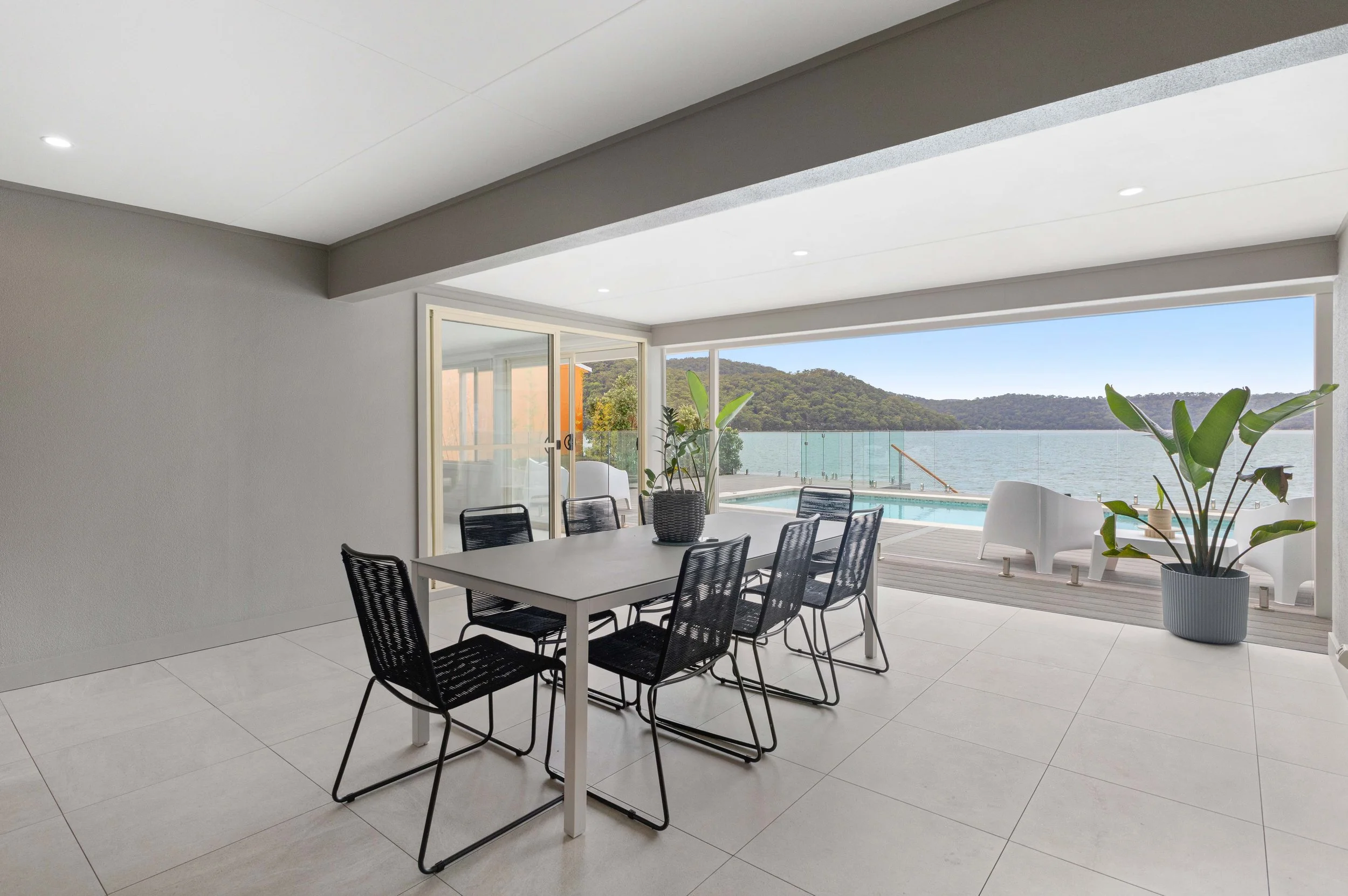 Modern dining room with a large table surrounded by black mesh chairs, opening to an outdoor deck with patio furniture and a view of a lake and green hills.