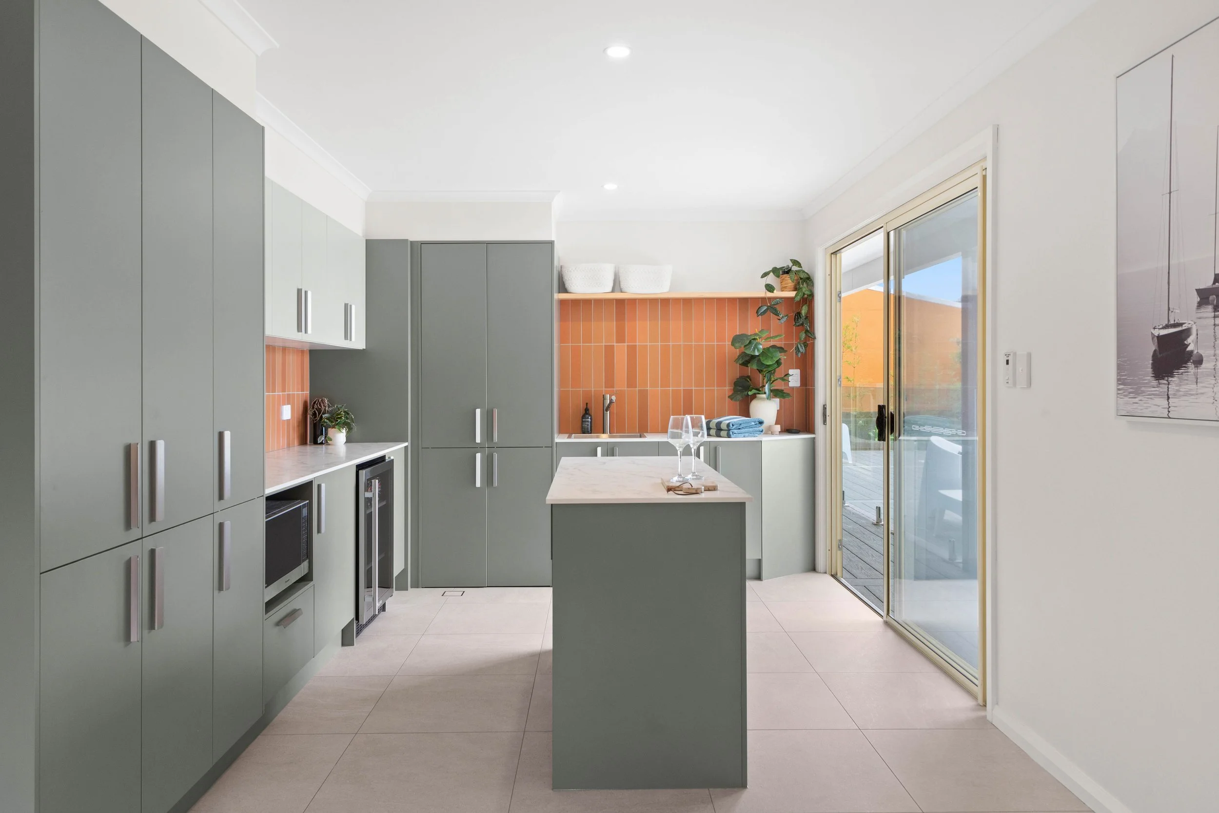 Modern kitchen with green cabinets, orange tiled backsplash, white countertops, and sliding glass door leading to outdoor patio. Decor includes potted plants, wine glasses, and books.