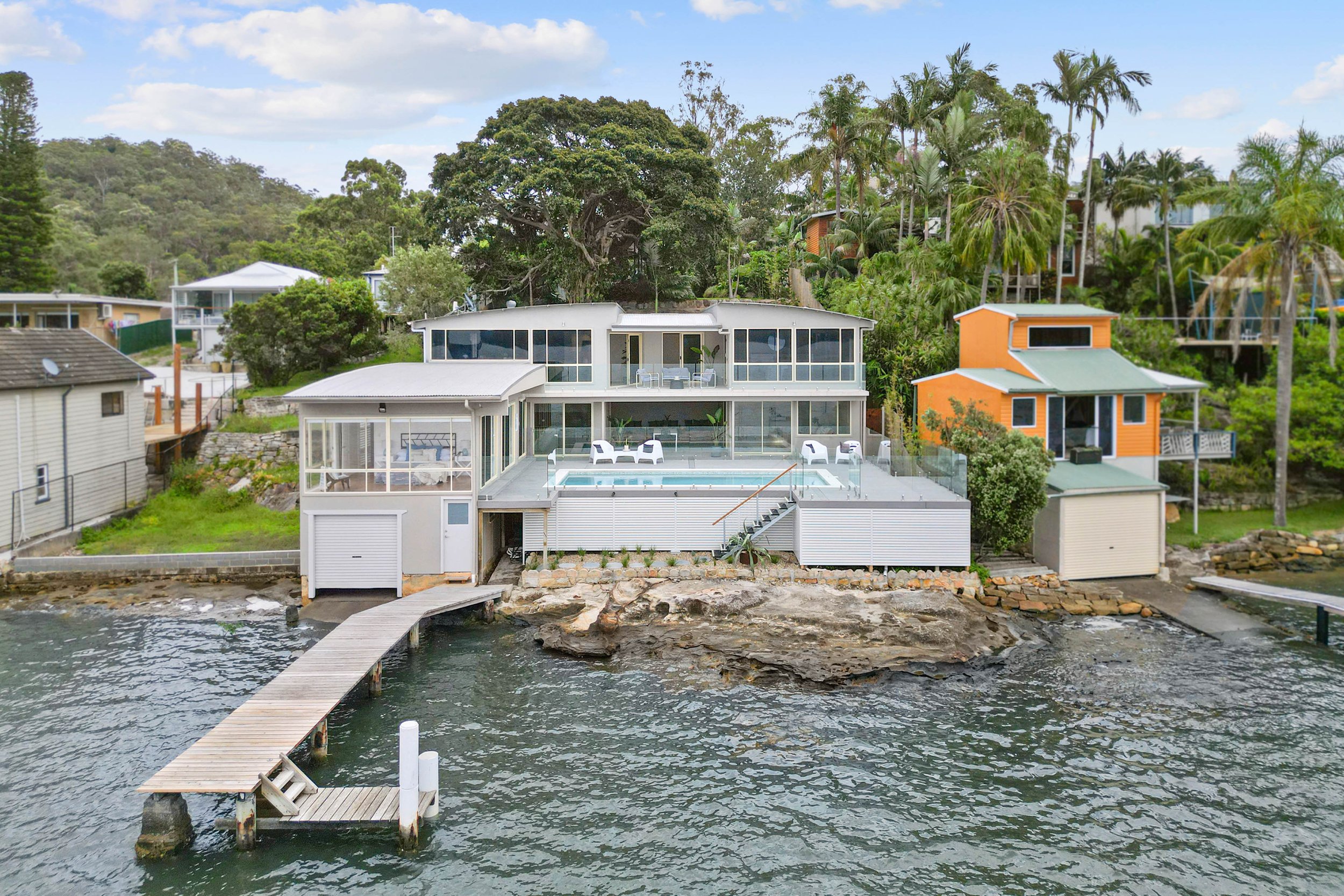 Modern house overlooking water with a wooden dock and stairs, surrounded by greenery and neighboring houses.