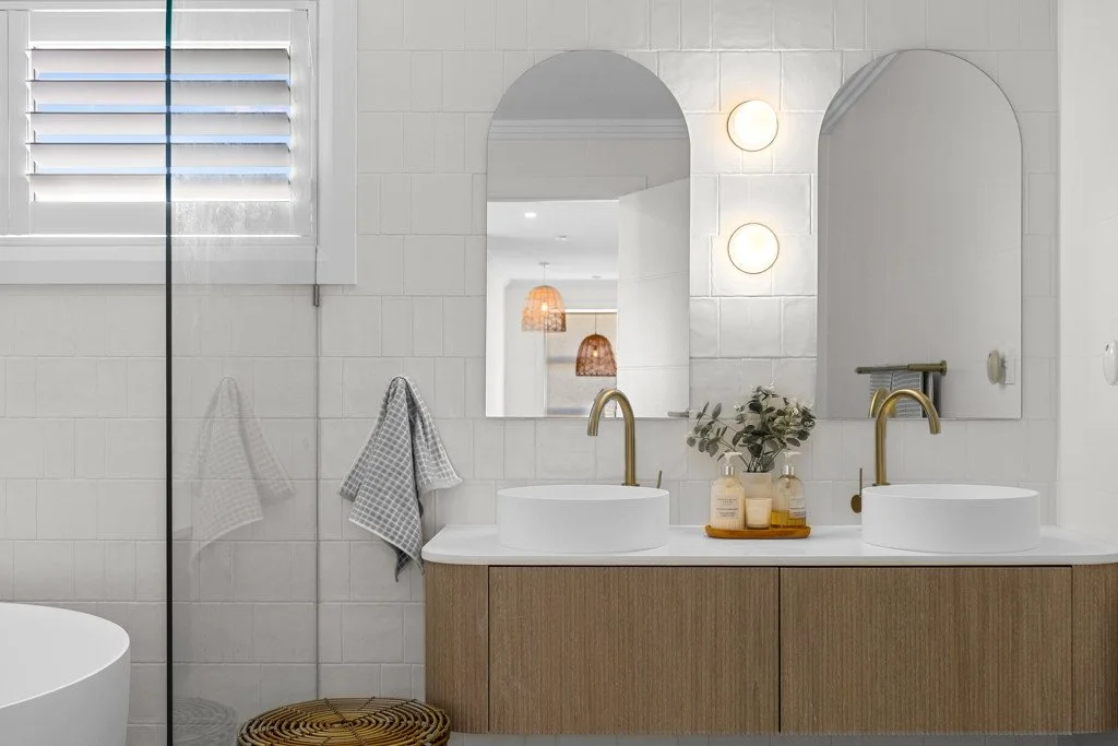 Modern bathroom with two round white sinks, gold faucets, large mirrors, a window with shutters, and decorative soap bottles on a wooden vanity.