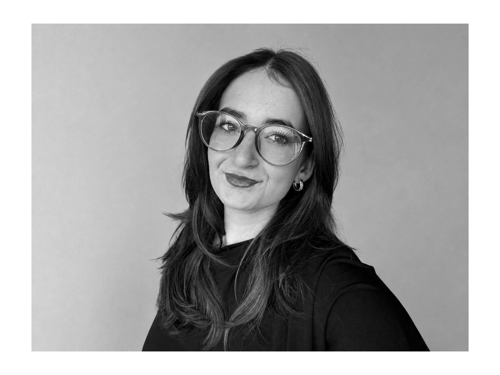 A black-and-white portrait of a woman with long wavy hair, wearing glasses, earrings, and a dark top, smiling slightly against a plain background.