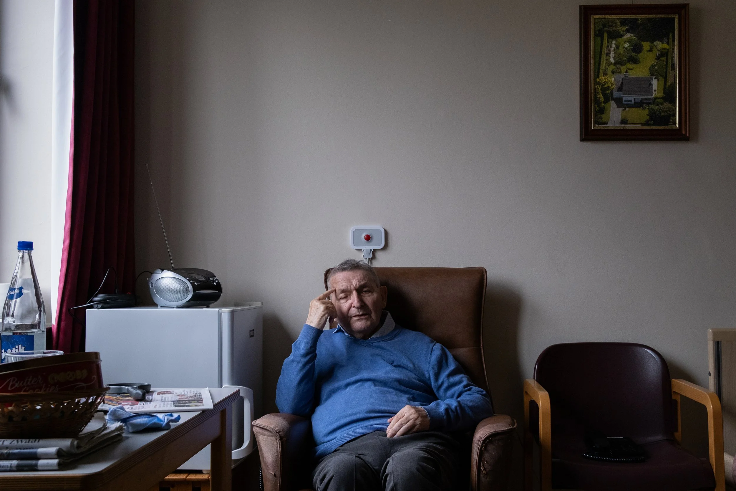 An elderly man sitting in a brown chair in a room with a white wall, a framed painting of a house and garden, a small refrigerator, a radio, a water bottle, and a table with newspapers and magazines. The man is wearing a blue sweater and appears to be scratching his head.