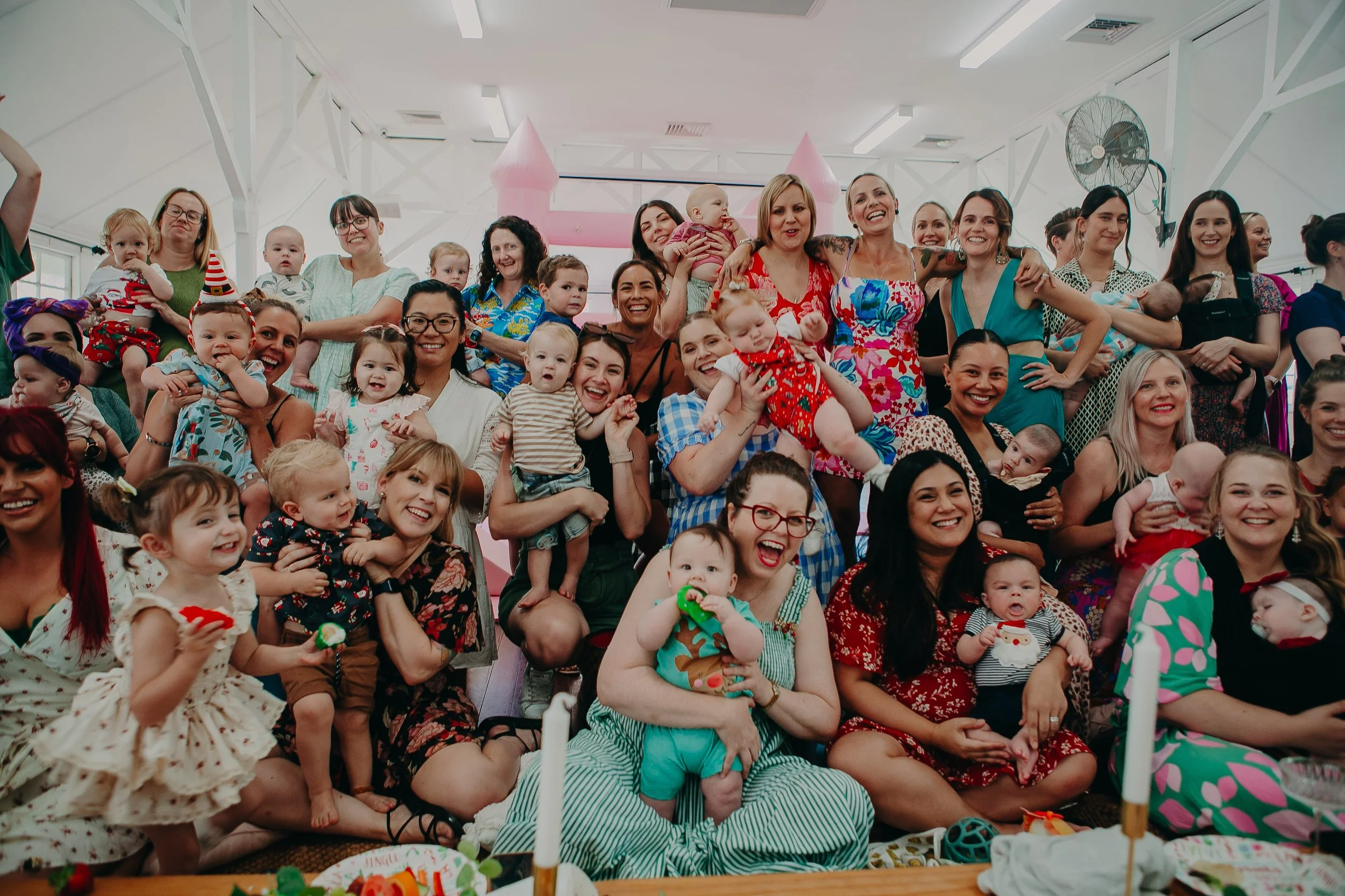 Group of women and children at a holiday celebration, smiling and posing indoors with decorations and a pink castle in the background.