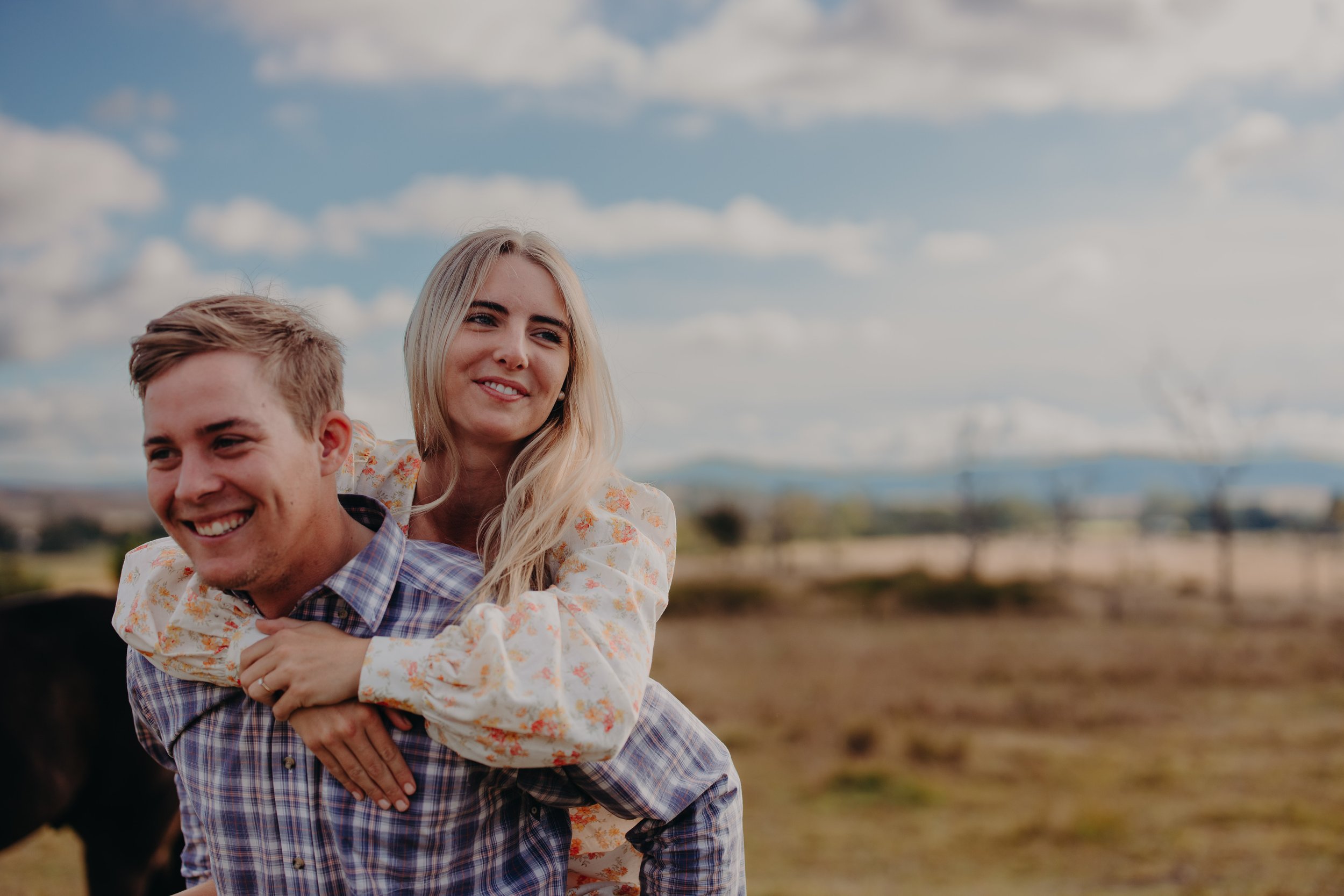 A young man giving a piggyback ride to a young woman in an open field with a blue sky and clouds.