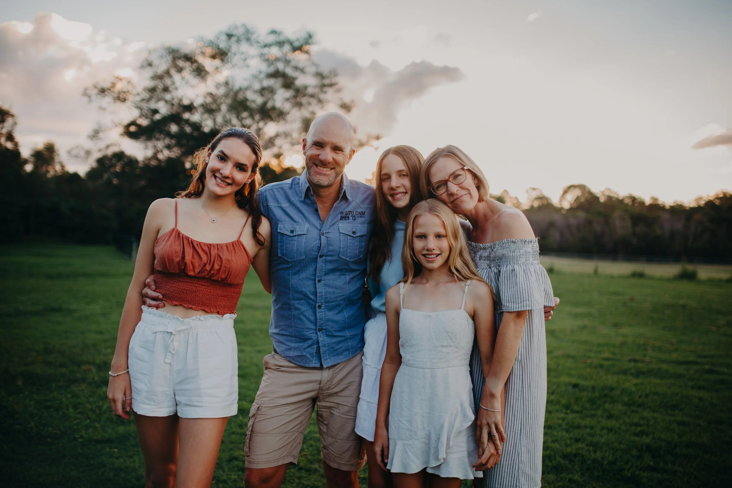 A group of six people, including children and adults, smiling and standing close together outdoors on a grassy field during sunset, with trees and a partly cloudy sky in the background.