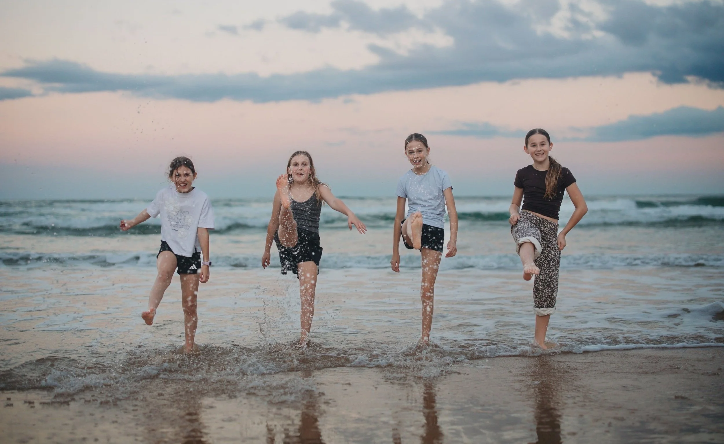 Four young girls playing and running in the ocean at the beach during sunset, splashing water, smiling, wearing casual summer clothes.