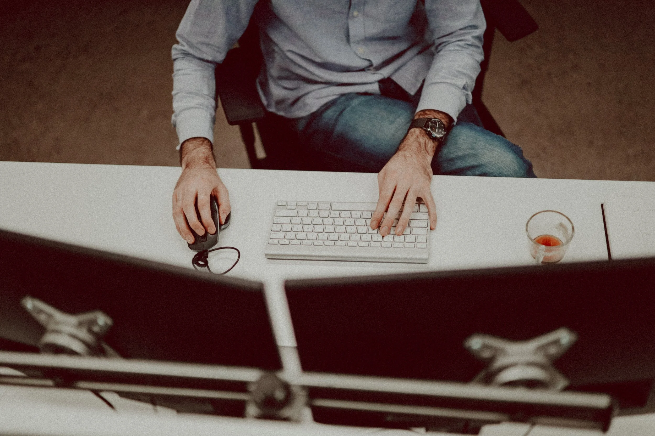A person working at a desk with a computer, mouse, keyboard, and a partially filled glass of tea.