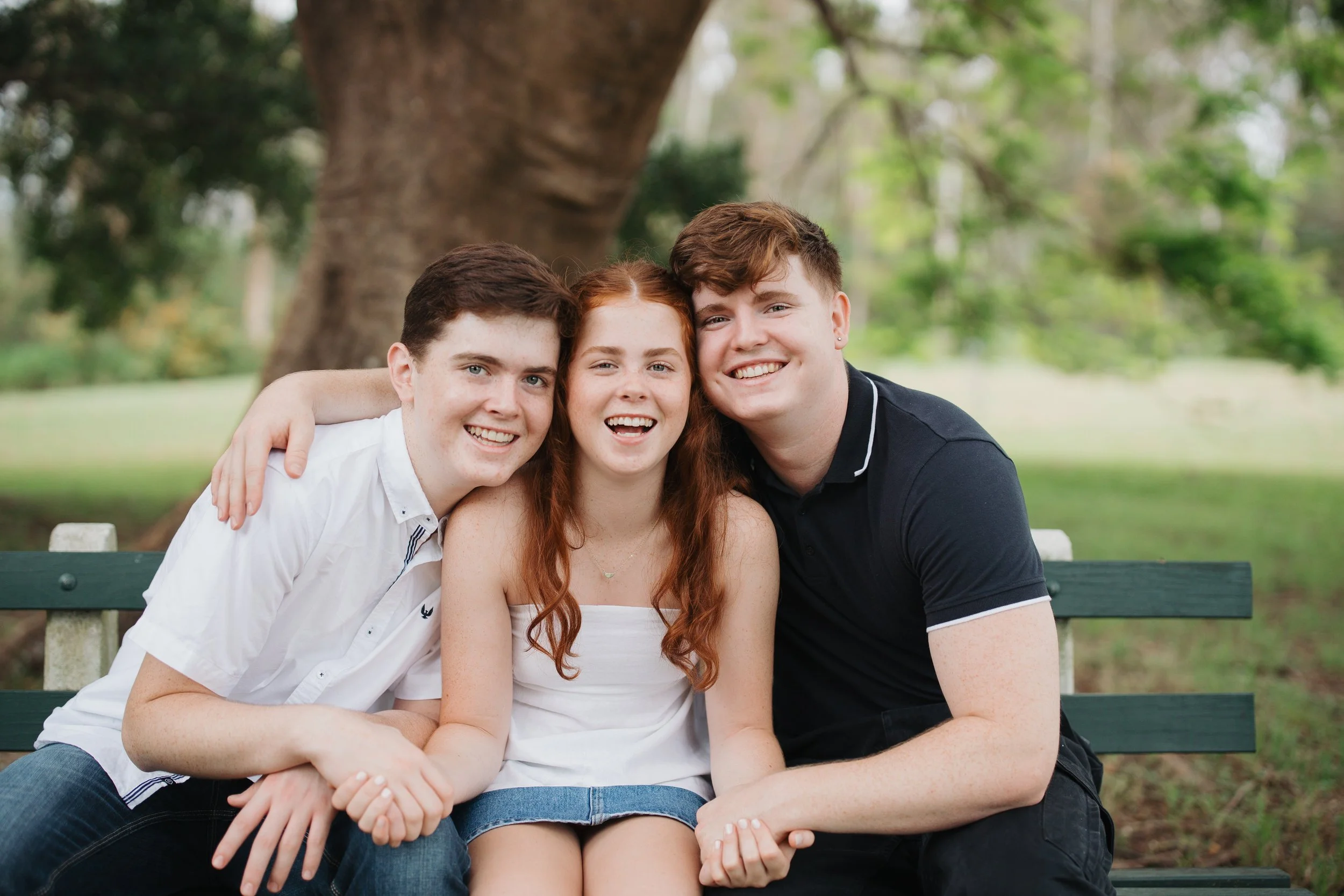 Three friends sitting on a green park bench smiling, with trees in the background.