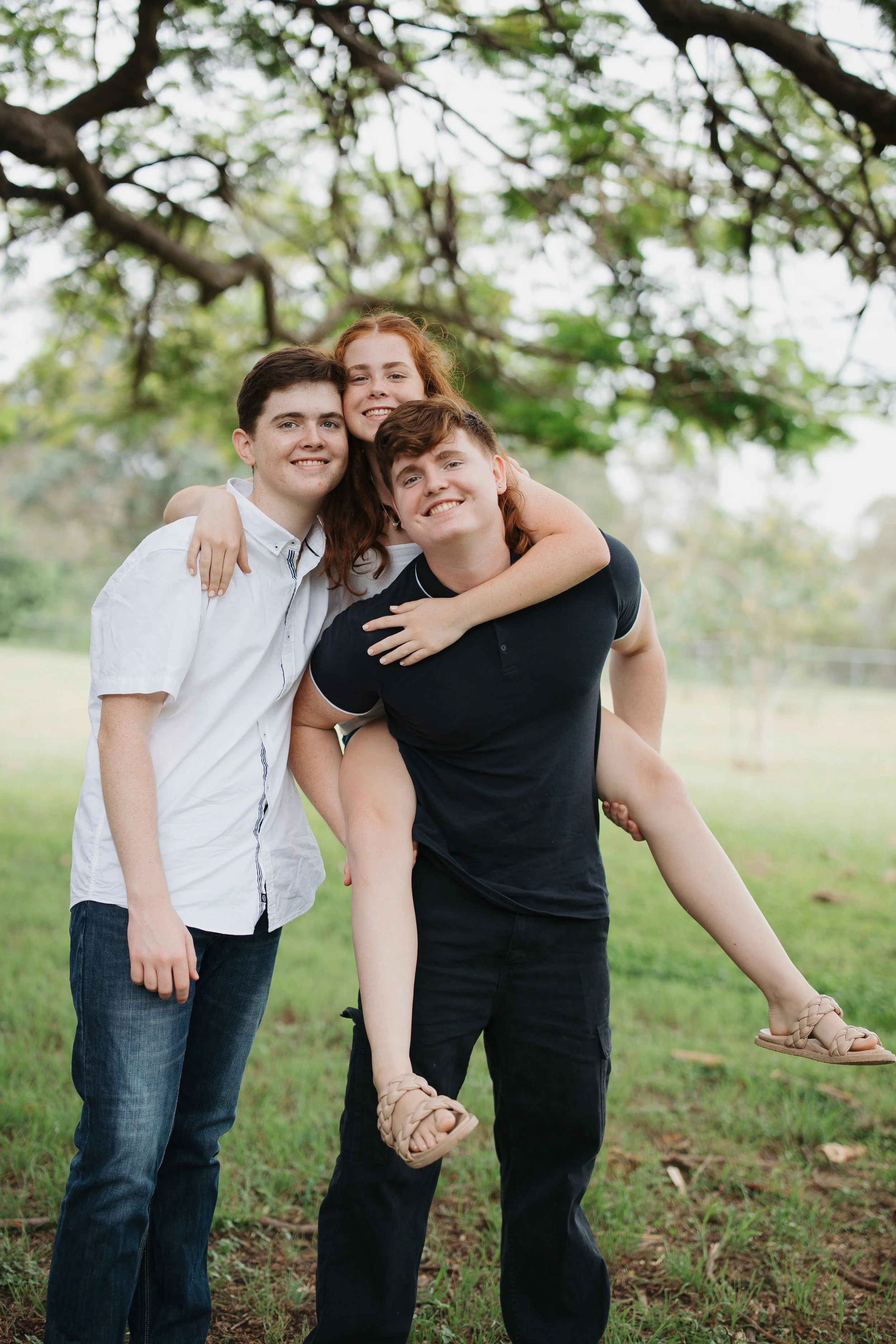 Three young people outdoors under a large tree with green leaves, smiling, with one person piggyback riding another.