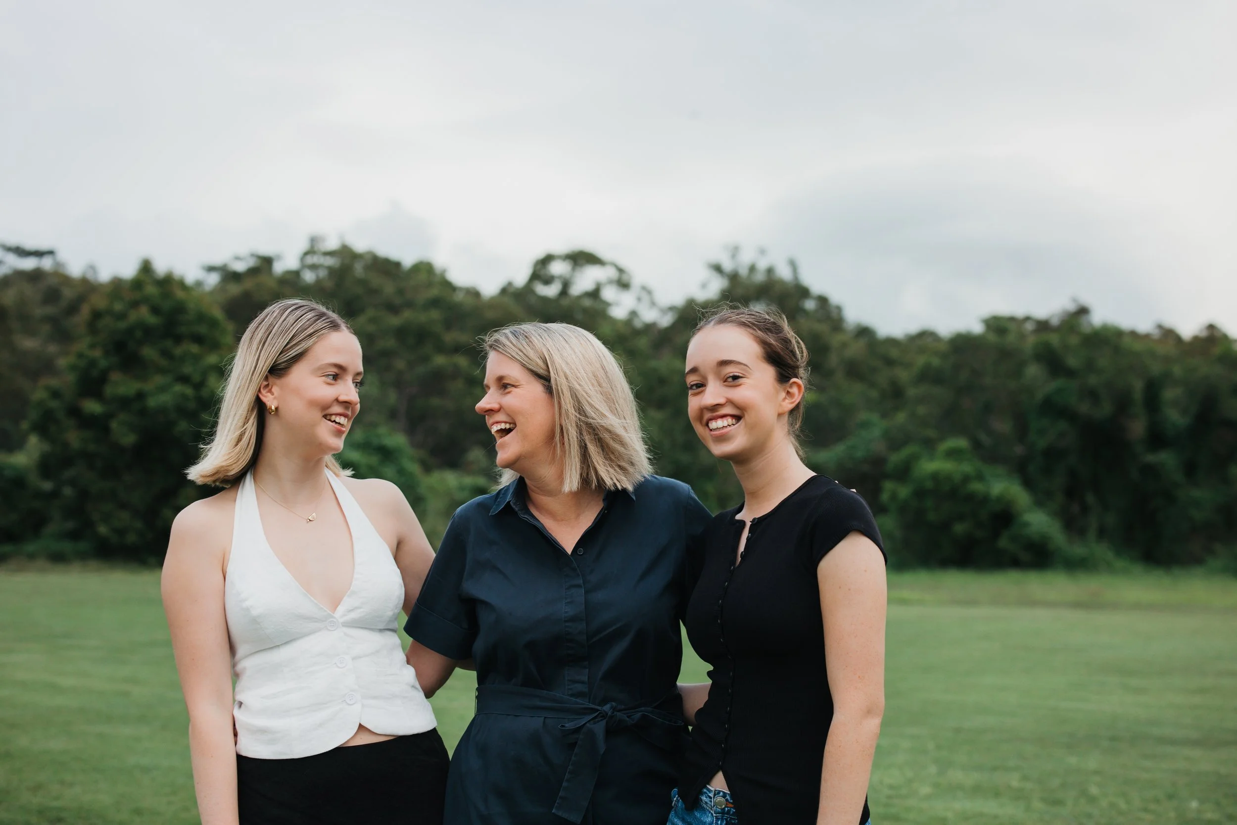 Three women standing outdoors on a grassy field, smiling and talking, with trees in the background.