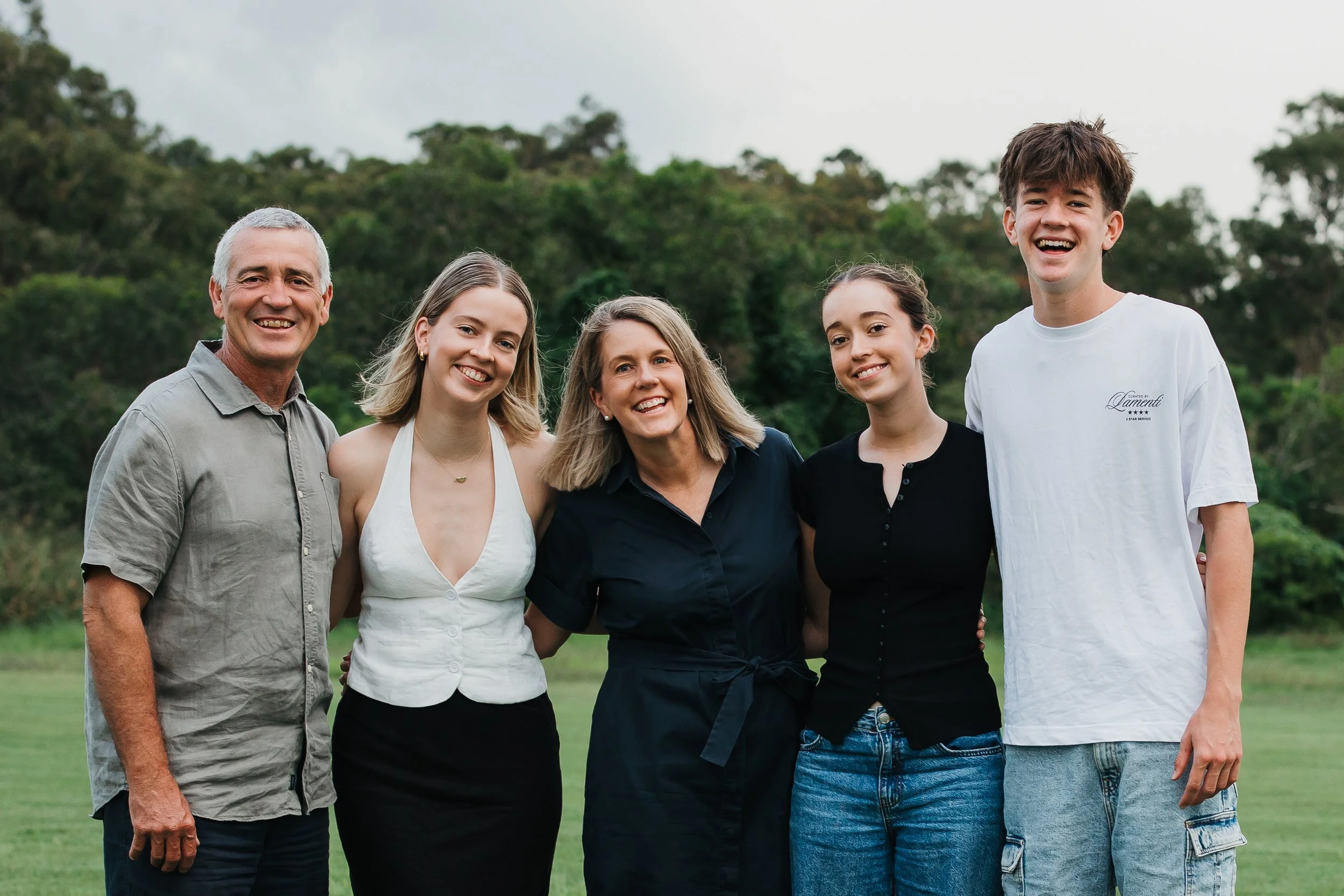 Group of five smiling people outdoors, standing in a line, with green trees in the background.